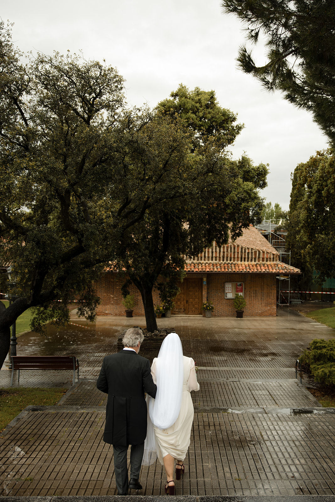Novia entrando a la iglesia con su padrino el día de su boda tras la lluvia, suelo mojado reflejando el momento